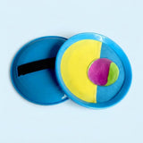 A catch game set with blue paddles, yellow-and-blue cloth faces, a black strap and a purple-and-green ball sits against a pale blue background.