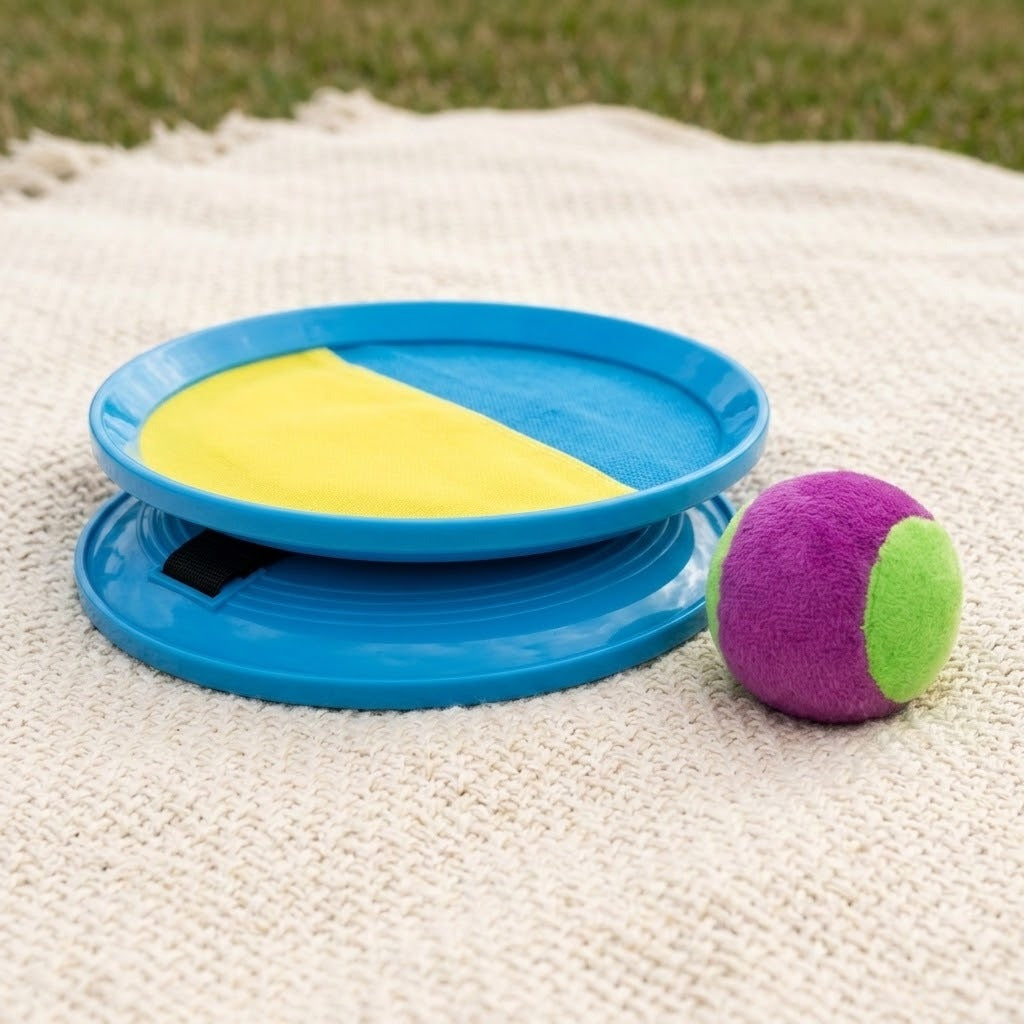 A catch game set with stacked blue paddles and a purple-and-green ball rests on a cream woven blanket outdoors.