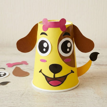 Paper cup craft kit shown assembled as a yellow dog with brown ears and pink accents beside loose cutouts on pale wood.