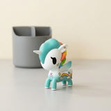 A white and turquoise unicorn-shaped blind box figure stands on a pale desktop beside a grey organizer.