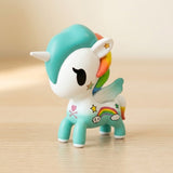 A white and turquoise blind box figure with rainbow details and small wings stands on a light wooden surface.
