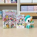A Tokidoki Unicorno Bambino Series 1 blind box figure is displayed next to its packaging on a light wooden tabletop. The figure is white with a teal mane and tail, and features a rainbow-colored horn and a small rainbow chest emblem.