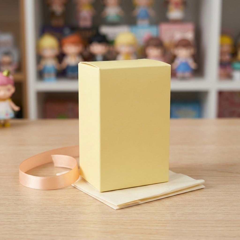 Blind box figure packaging in pale yellow rests on cream paper beside peach ribbon on a wooden table, with blurred figures behind.