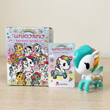A white and turquoise blind box figure stands beside one illustrated box and one accompanying card on a wooden desktop.