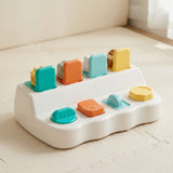 White pop-up toy with four colorful controls and upright lids, viewed at an angle on a cream play mat.