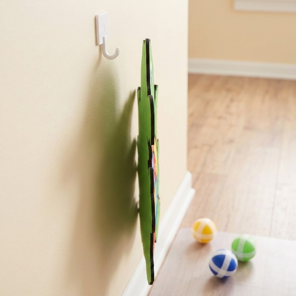 Green sticky ball target viewed edge-on against a beige wall, with a white hook nearby and three colored balls on the wooden floor.