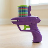 Purple plastic disc shooter with a green trigger and clear chamber of colorful discs stands on pale wood near a window.