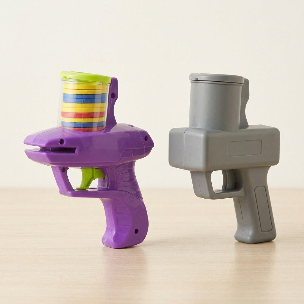 A purple plastic disc shooter with colorful discs stands beside a gray disc shooter on light wood against a cream background.