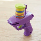 Purple plastic disc shooter viewed from above stands on light wood with colorful discs beneath its closed green lid.