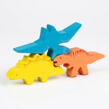 Three wooden dinosaur stacking blocks balance against a white background, with a blue winged piece above yellow and orange dinosaurs.