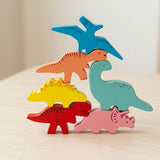 Six colorful wooden dinosaur stacking blocks balance in a tower on a light wood surface against a soft beige background.