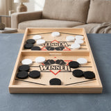 Wooden sling puck game with black and white pucks and a central opening on a gray tabletop before a beige sofa.