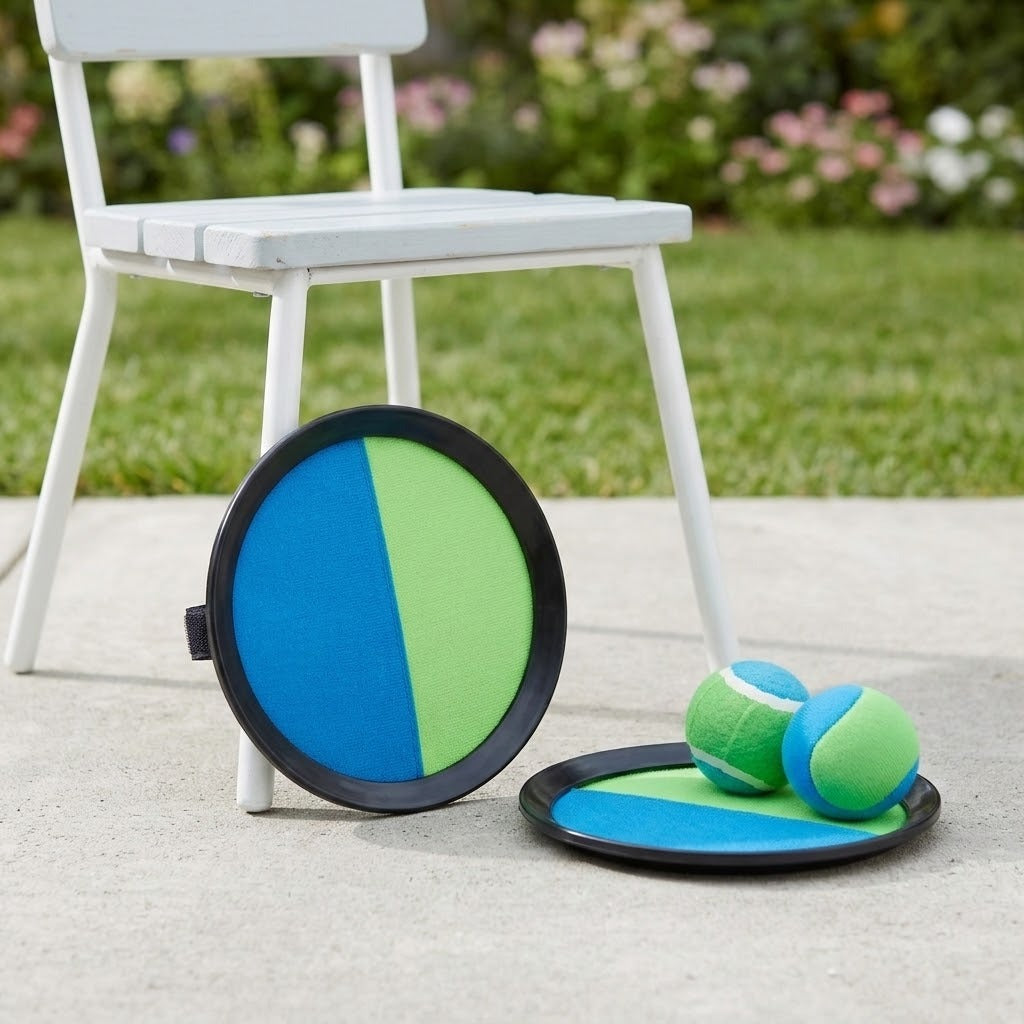 Blue and green catch game set with two black-rimmed discs and two balls rests beside a white chair on a garden patio.