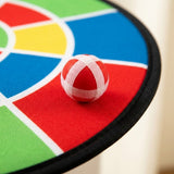 A red ball with white gripping strips rests on the textured, multicolored cloth dart board beside its black edging.
