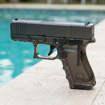 Dark gray water gun with a translucent textured grip stands on pale stone paving beside a turquoise swimming pool.
