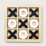 A wooden 3x3 tic-tac-toe game board with black X shapes and white O shapes arranged on a light natural wood frame, photographed from above on a plain light background.