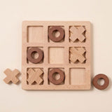 A wooden tic-tac-toe game board with a natural light wood finish, featuring carved squares and circular holes. Two dark brown wooden pieces are placed on the board, and two additional loose pieces sit nearby on a warm cream background.