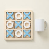 Wooden tic-tac-toe game with blue X pieces and white O pieces beside a white mug on a cream background.