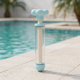 Light blue water gun with a clear barrel and cream inner shaft stands upright on paving beside a swimming pool.