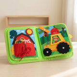Green fabric busy book propped open on a light wooden surface, showing a red barn pocket and yellow tractor activity.
