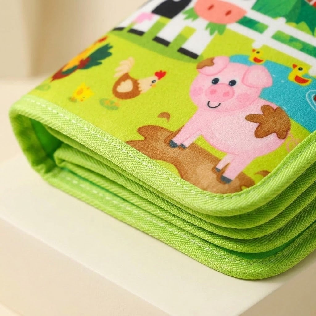 Close-up of a green fabric busy book showing stitched edging, layered pages, and a pink pig illustration on a light surface.