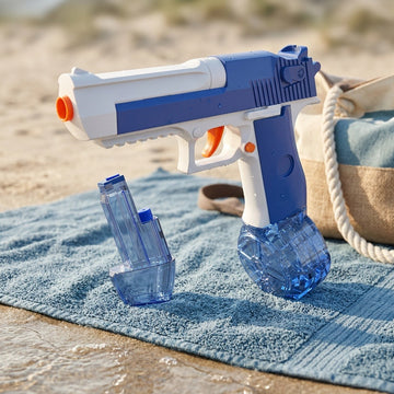 Blue-and-white plastic water gun with orange trigger and two transparent reservoirs on a beach towel near a woven bag.
