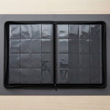 Open black trading-card binder with empty clear pocket pages lies on a dark gray mat over a pale wooden desk.