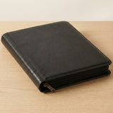 Closed black zippered trading card binder lying flat on a light wooden table with the zipper pull visible.
