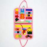 Pink felt busy board displayed open against a white background, with carrying loops, colorful fastening activities and felt shapes.
