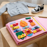 Pink felt busy board on a wooden stool, showing buckles, buttons and shoelace activities, with a cardigan and sneaker behind.