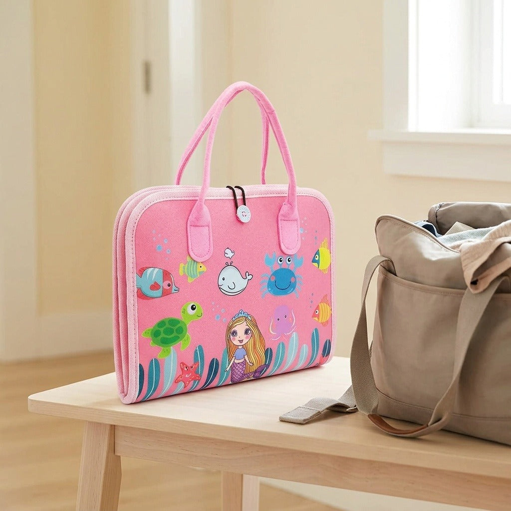 Closed pink busy board with a mermaid and sea creature cover stands on a wooden bench beside a beige bag.