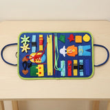 Blue felt busy board lies open on a light wood table, displaying colorful buckles, zippers, laces and shapes with green edging.