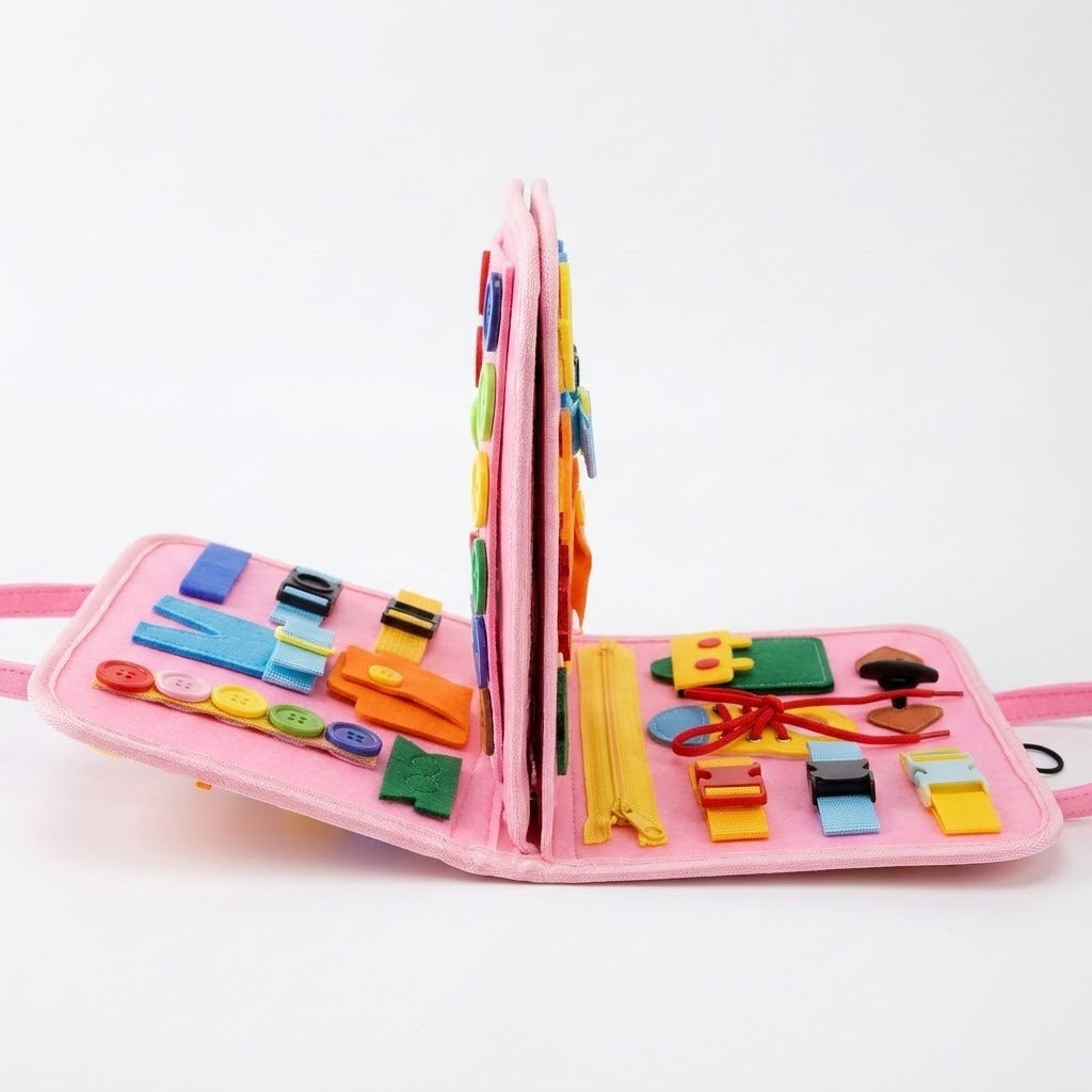 Pink felt busy board stands open with upright central pages, colorful buckles, buttons and a yellow zipper against a white background.