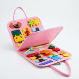 Pink felt busy board sits partly open on a white background, showing a raised central page, colorful shapes, buckles and laces.