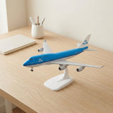 A blue and white aircraft model sits on a white display stand on a wooden desk beside a notebook and pencil cup.