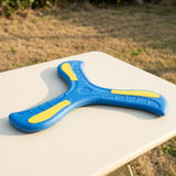 Blue and yellow three-arm boomerang lying flat on a white outdoor table with blurred grass behind.