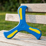 Blue and yellow three-arm boomerang propped against a weathered wooden bench with blurred greenery behind.