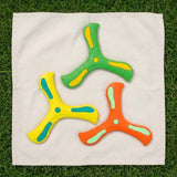 Three boomerangs in green, yellow and orange with contrasting accents arranged on a cream cloth over grass.