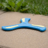A blue three-blade EVA boomerang with pale yellow accents rests on concrete with blurred green grass behind it.