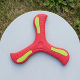 A red three-blade EVA boomerang with yellow-green accents rests on a white circular surface outdoors beside grass.