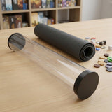 A transparent playmat storage tube with black end caps rests on a wooden table beside a rolled dark gray mat and game pieces.