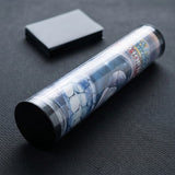 Transparent playmat storage tube with black end caps holds a printed rolled mat beside a rectangular box on a dark textured surface.