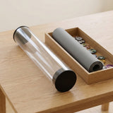 A transparent playmat storage tube with black end caps rests beside a wooden tray holding a rolled gray mat and game tokens.
