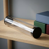 Empty transparent playmat storage tube with black end caps lies on a wooden shelf beside blue, green, and red storage boxes.