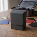 A dark gray textured deck box with a stitched flap sits on a wooden table among cards, with an open carrying case behind it.