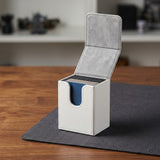 An open white deck box holding cards reveals gray lining on a dark gray mat atop a wooden table.