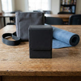 A black textured deck box with a closed stitched flap sits on a wooden table beside a gray bag and rolled blue mat.