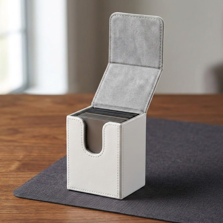 A light gray faux-leather card deck box with an open hinged lid, standing on a dark gray mat on a wooden table.