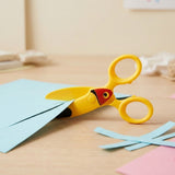 Yellow bird-design safety scissors shown cutting light blue paper on a wooden craft table beside paper strips.