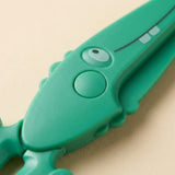 Close-up of green crocodile-design safety scissors showing the circular pivot, textured surface, and printed eye against a beige background.